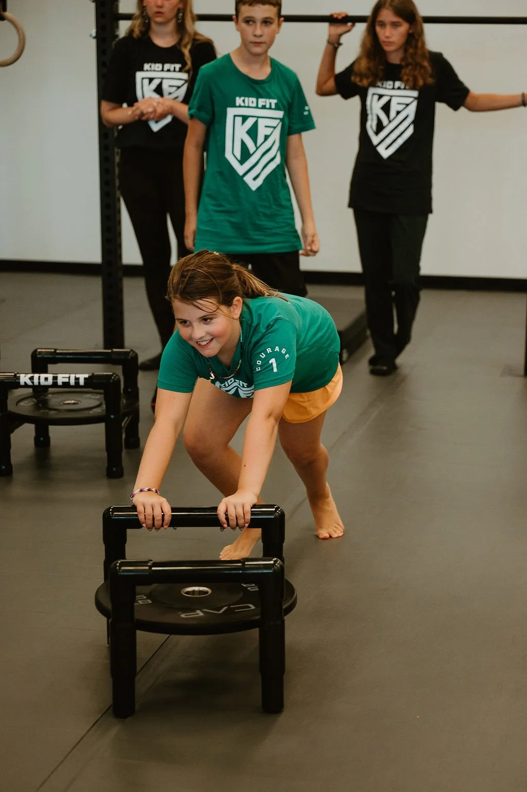 A young girl is playing a fitness game on a small black step platform in a gym, with other children observing in the background.