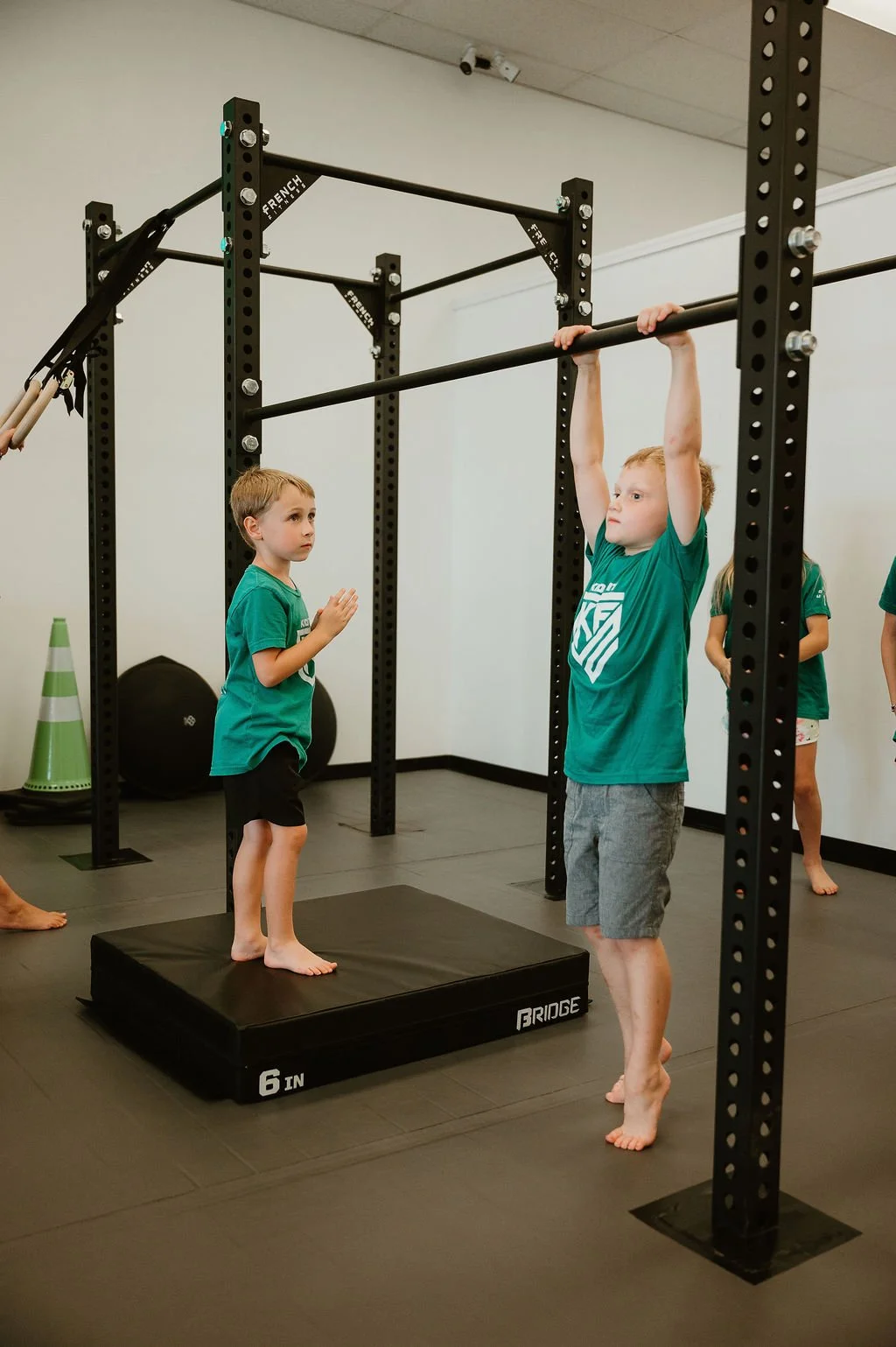 Young children in a gym practicing exercises with a pull-up bar and a mat, while an instructor observes.