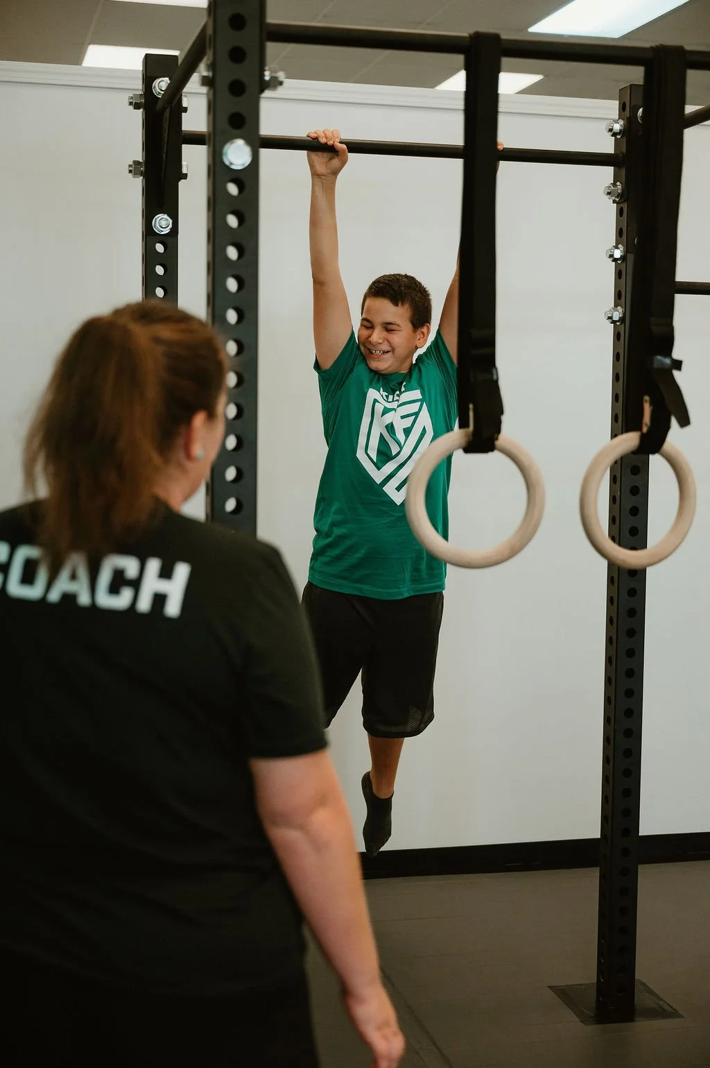 A young boy hanging from gymnastic rings during a fitness training session, supervised by a coach.