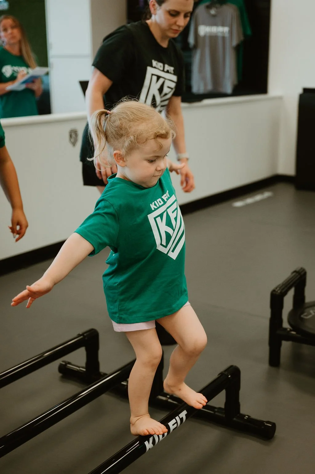 A young girl with blonde hair in a ponytail balancing on a black KidFit agility ladder during a fitness class, with a woman in a black KidFit shirt overseeing the activity.