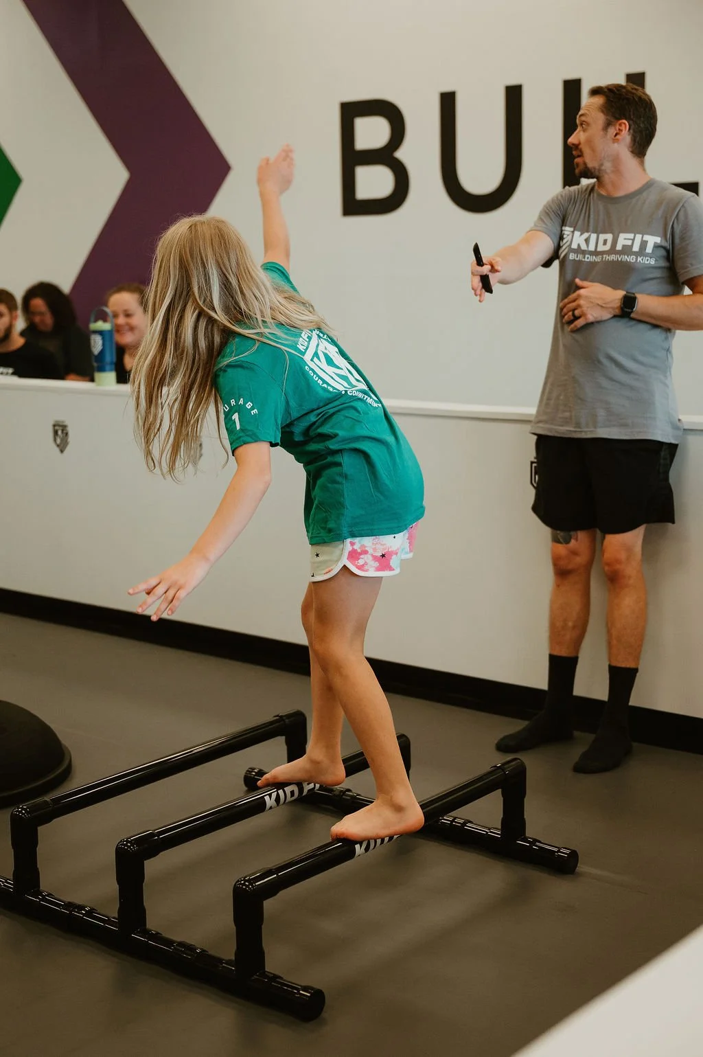 A young girl balancing on a small black platform in a fitness or training facility. An instructor nearby is giving instructions, with a group of people seated at a table in the background. The girl is wearing a green Kid Fit T-shirt and shorts.
