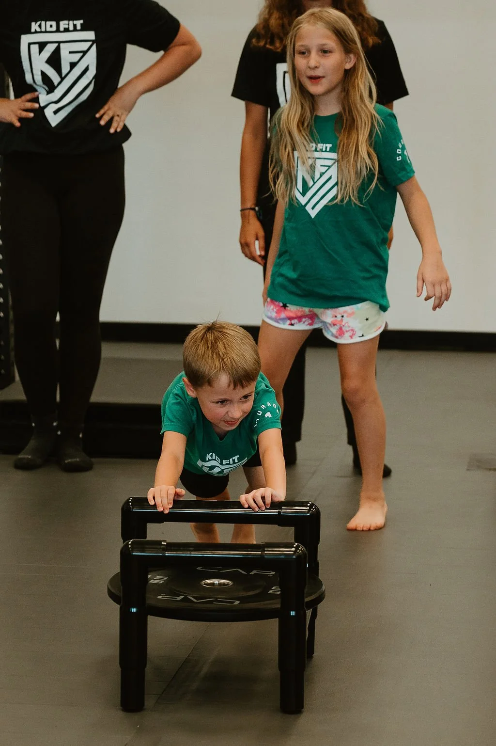 A young boy and girl playing on a black indoor fitness platform, with the boy pushing a sled, and the girl watching nearby. Two women stand behind them, observing.