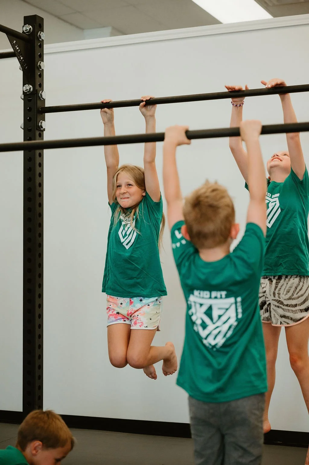 Children at a Kid Fit gym hanging from a horizontal pull-up bar, with one girl in mid-air smiling, wearing a green shirt and shorts.