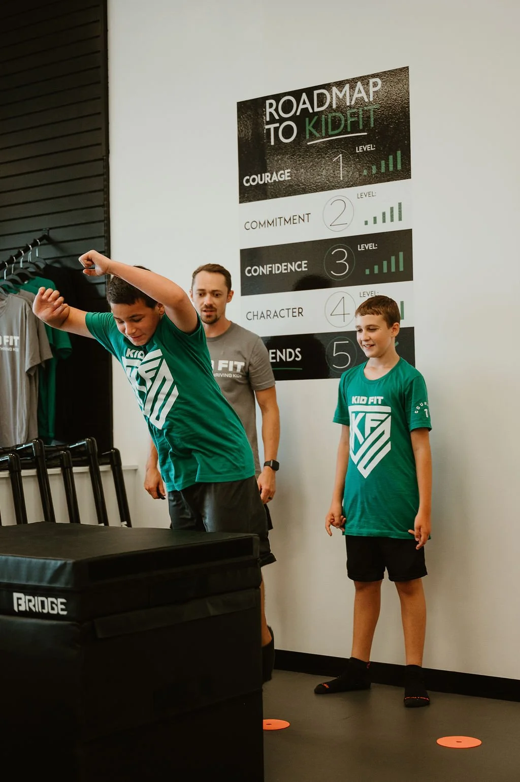 A young boy in a green 'Kid Fit' t-shirt jumping on to a training box while a man and another boy observe, standing near a large poster titled 'Roadmap to Kid Fit' with levels from 1 to 5.