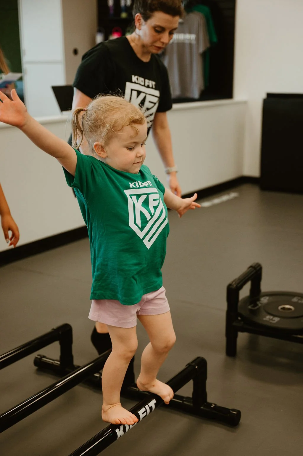 Young girl practicing balance on a black and metal balance beam during a fitness class, with an instructor standing behind her, observing.