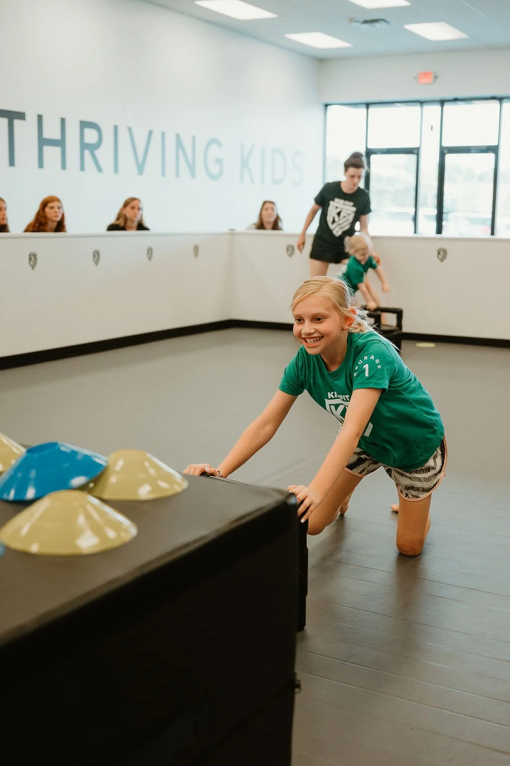 A young girl in a green Kid Fit shirt pushing a table with colorful bowls during a game or activity at a kids' program room, with other children and an instructor in the background, and the words 'THRIVING KIDS' on the wall.