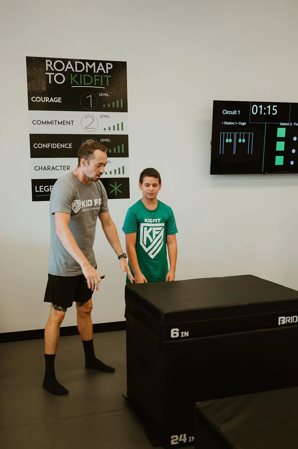 A man and a boy standing in a fitness or training facility near a foam padded box with a board and a motivational poster on the wall behind them.