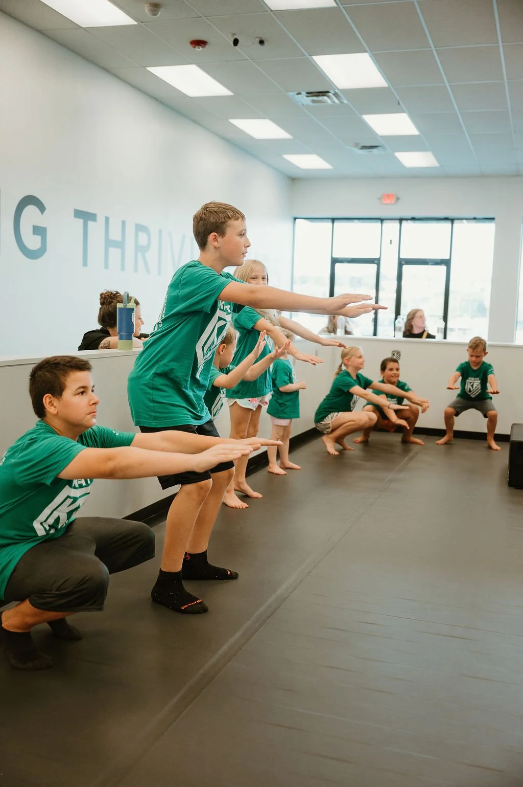 Children participating in a group exercise or dance class in a studio with large windows, wearing matching teal shirts.