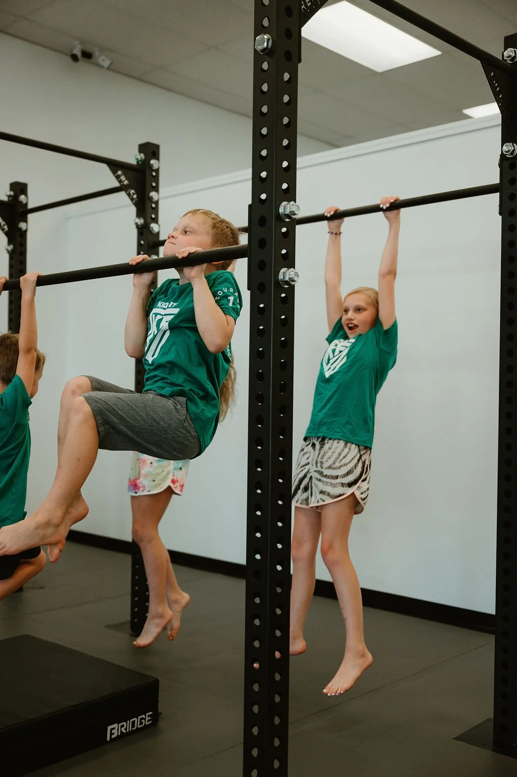 Children in green Kid Fit shirts hanging from gymnastic bars in a gym.