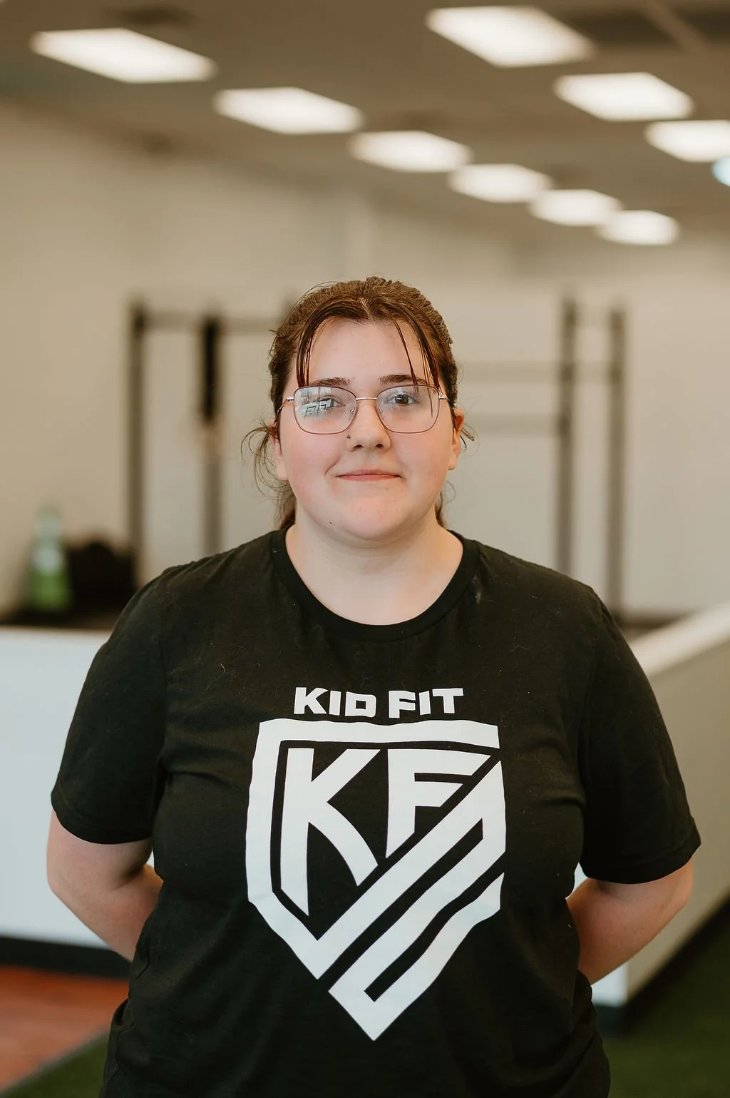 A young woman with glasses, wearing a black Kid Fit shirt, standing indoors with a neutral background.