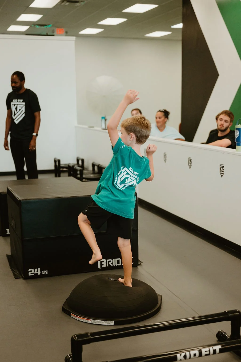 Young boy balancing on a black stability ball while raising one arm, in a gym with a padded black box and spectators watching.
