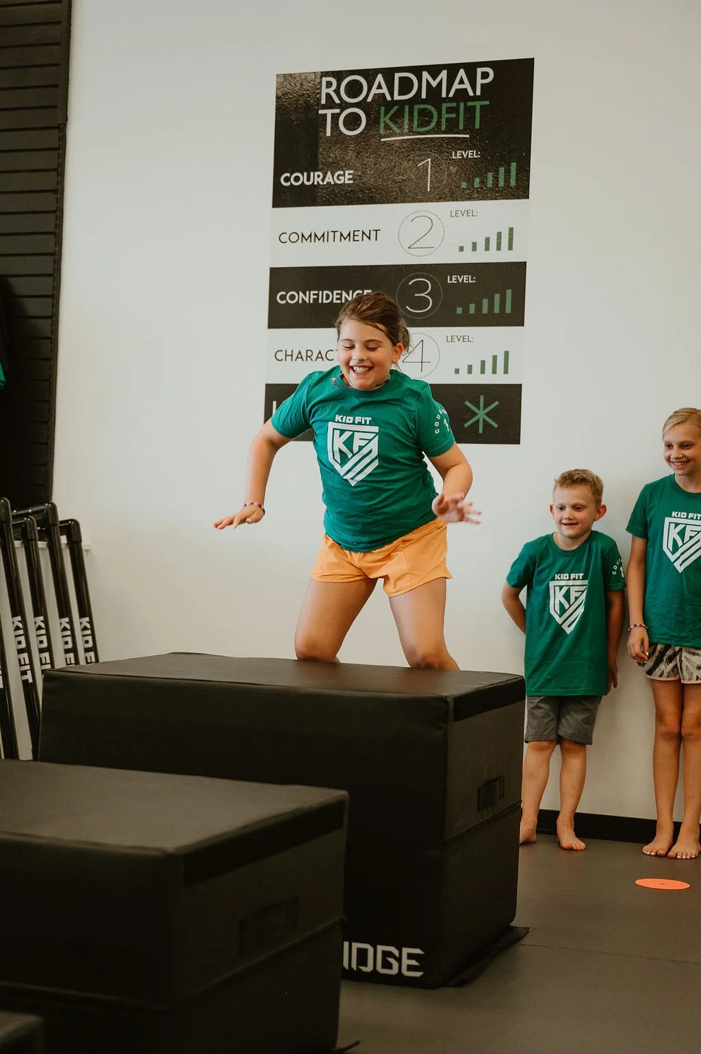 A young girl in a green Kid Fit T-shirt jumping onto a black padded box while smiling, with two children observing and smiling in the background inside a fitness room.