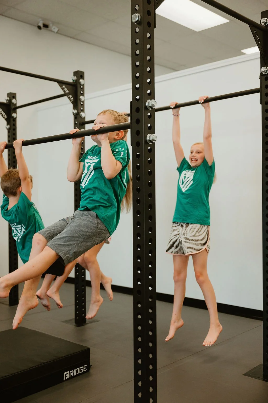 Children in a gym playing on horizontal bars, wearing green shirts and shorts, with some hanging and some smiling.