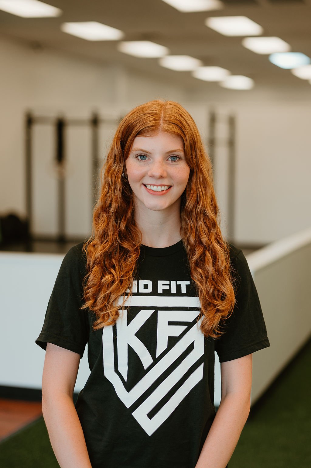 A young woman with long curly red hair smiling in an indoor gym, wearing a black T-shirt with white text and design that says "Kid Fit".