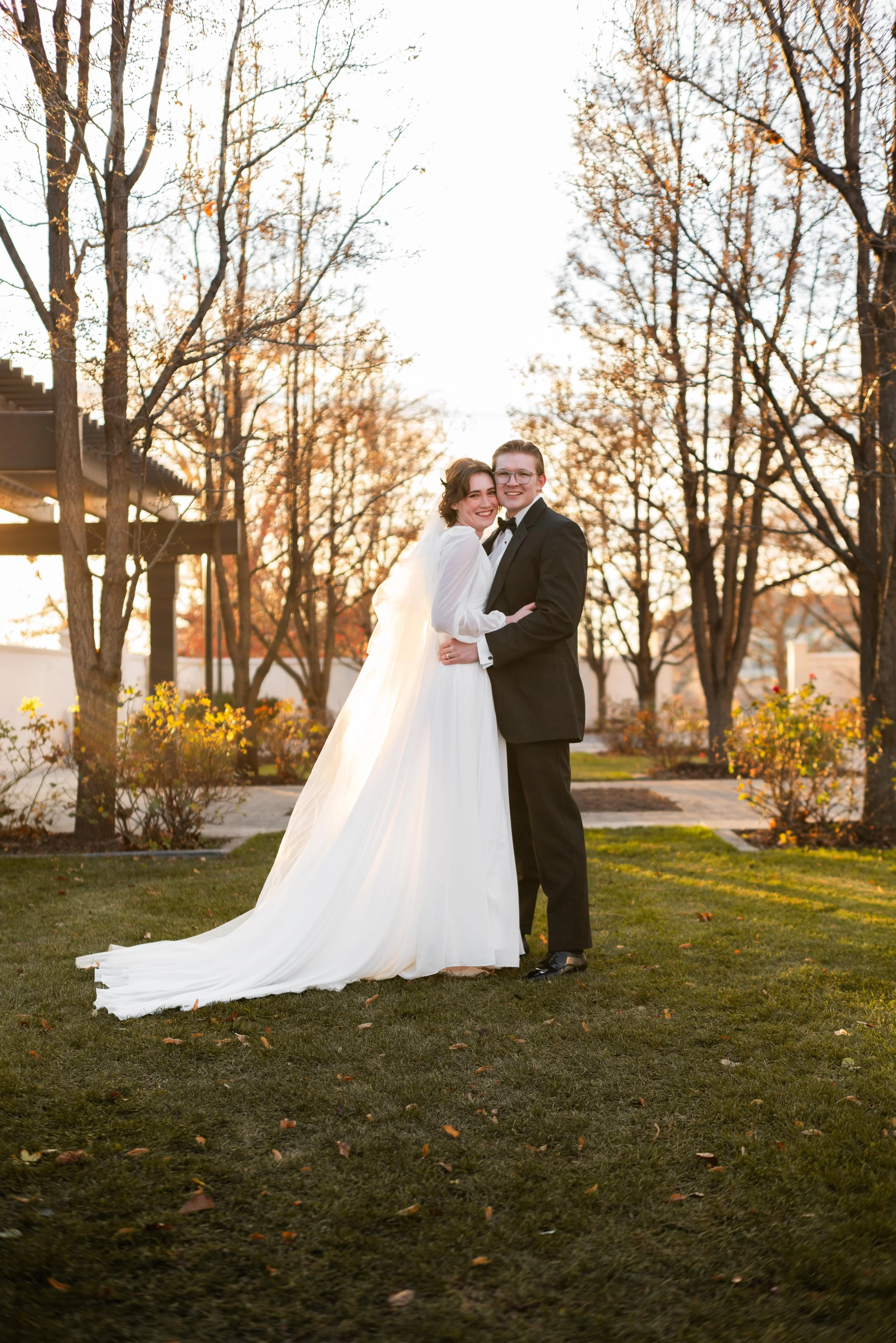 A bride and groom happily pose together outdoors during sunset, with trees and shrubs in the background, the bride wearing a white wedding gown and the groom in a black tuxedo.