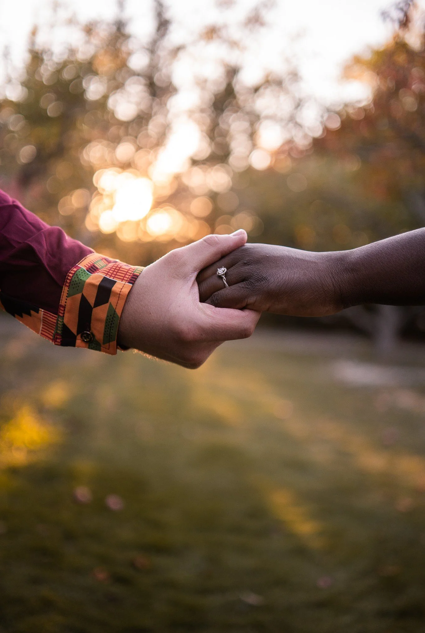 Two people holding hands outdoors at sunset, one wearing a ring on their finger.