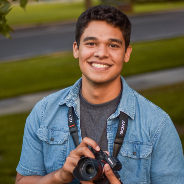 Young man smiling outdoors holding a Sony camera with a strap around his neck