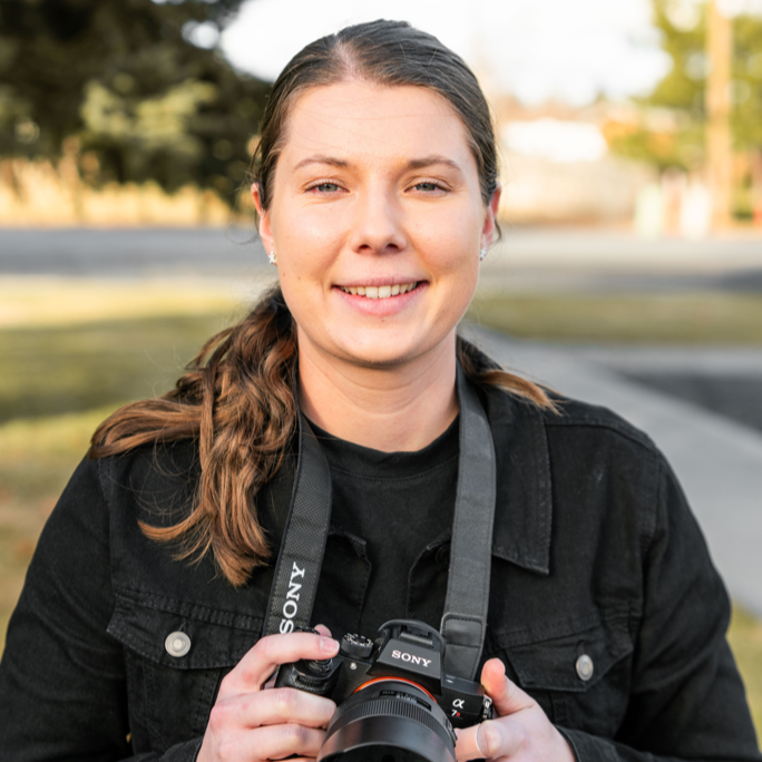 Young woman outdoors holding a camera and smiling at the camera.