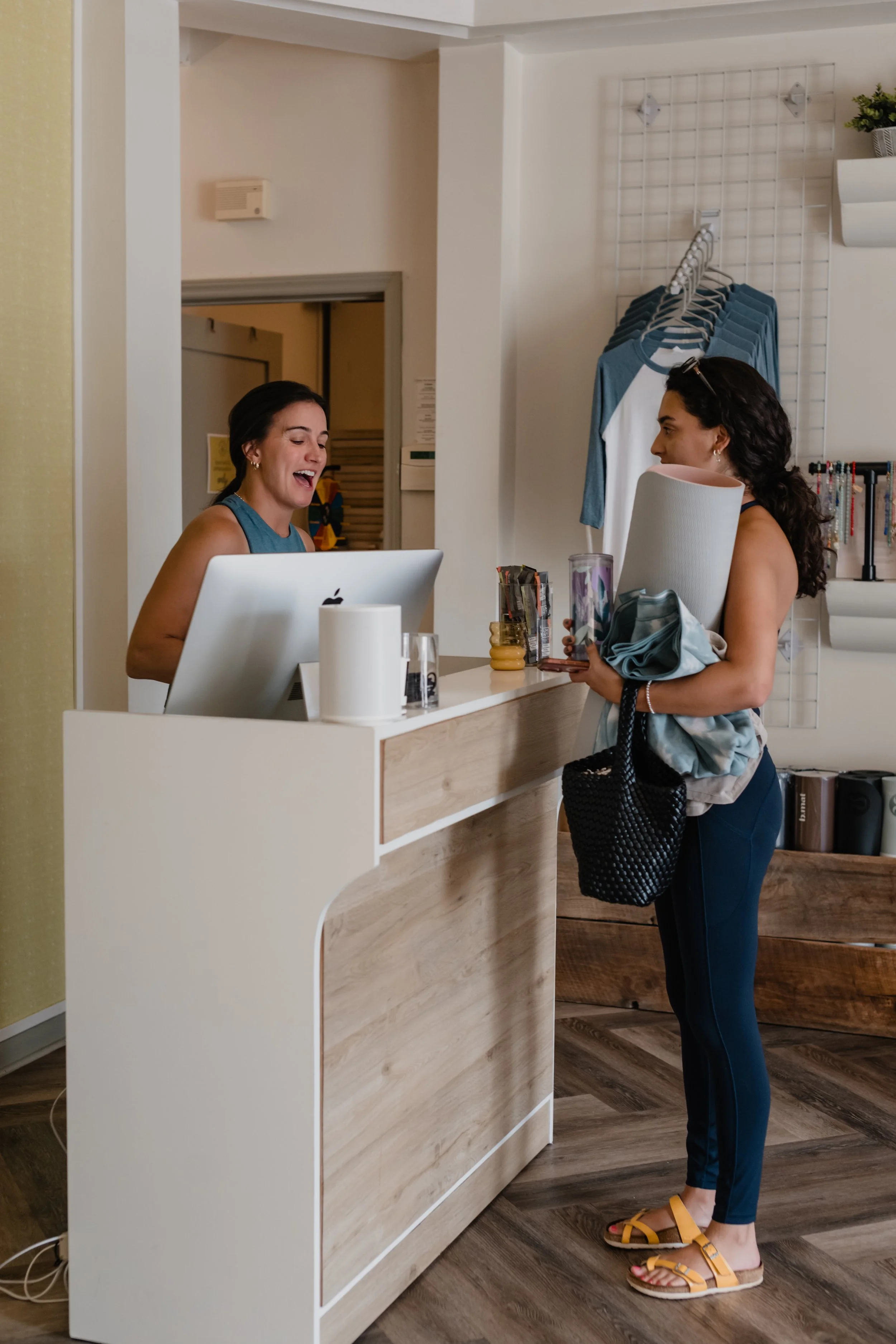 A woman is handing over a package and a rolled-up yoga mat to a receptionist at a store counter. The woman has dark curly hair and is wearing a navy blue outfit and yellow sandals. The receptionist, a woman with short dark hair, is smiling and has a computer and various items on the counter. In the background, there are blue T-shirts hanging on a rack and some wall decorations.