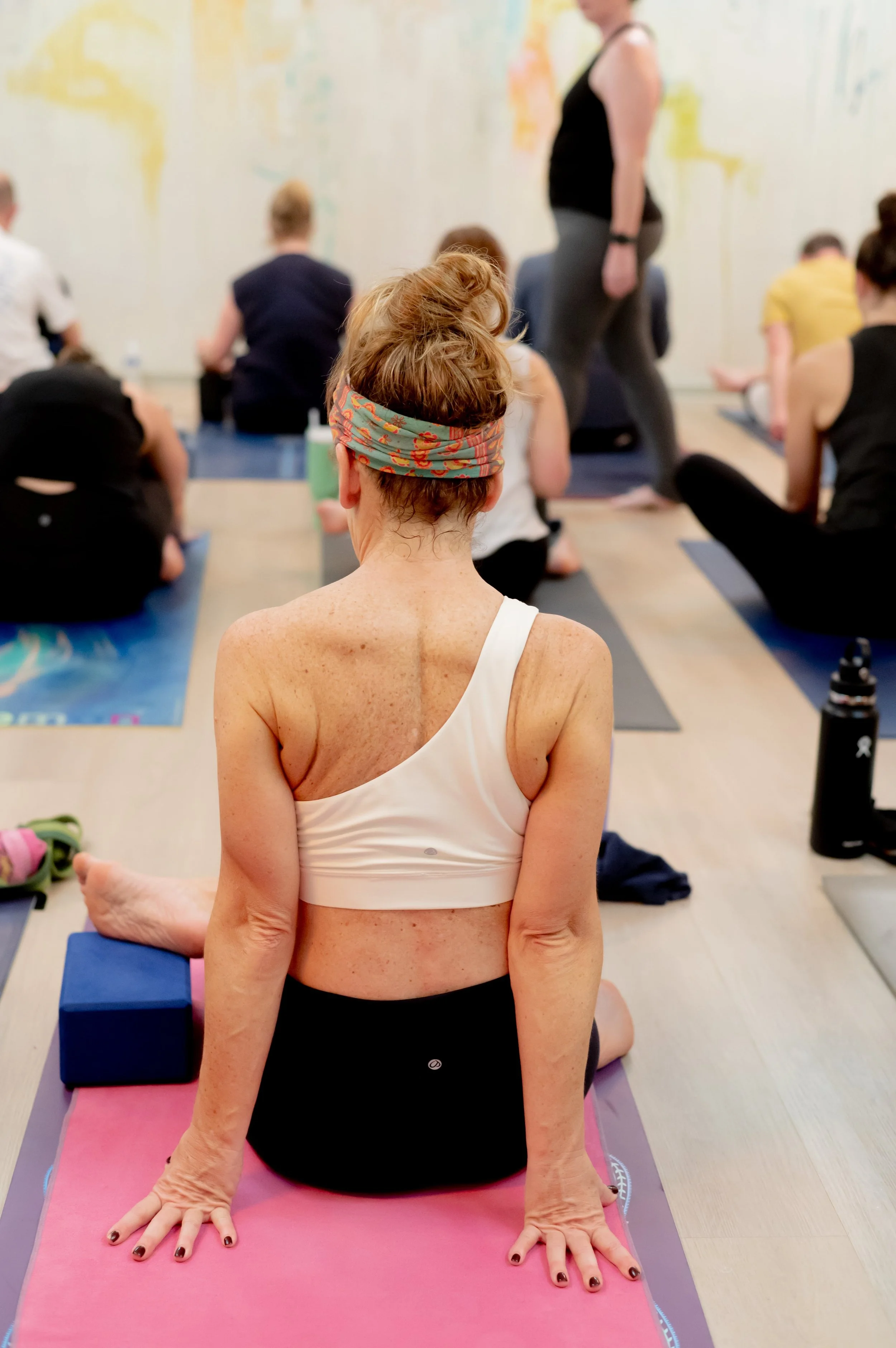 A woman with curly hair tied back, wearing a colorful headband and a white sports bra, practicing yoga in a group class with other participants. She is sitting on a pink yoga mat with her hands on the floor behind her, indoors with a colorful wall in
