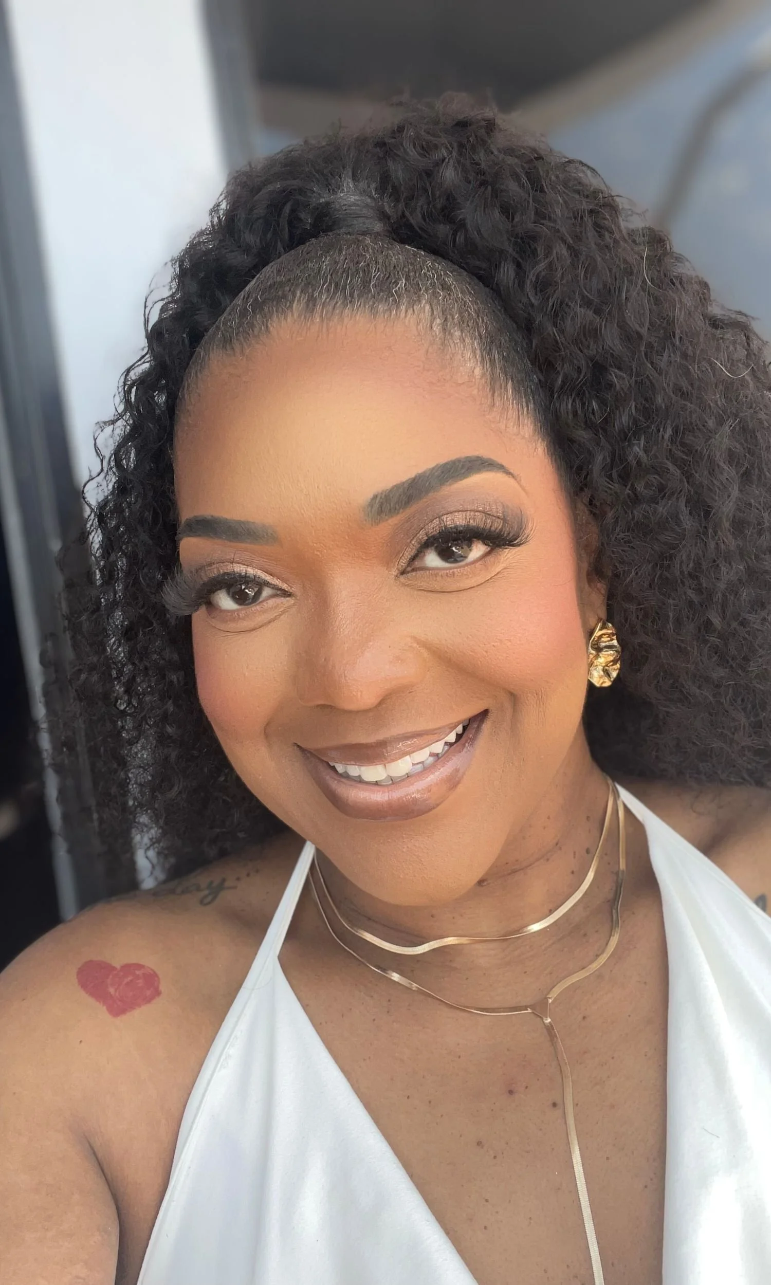 A woman with dark curly hair in a high ponytail, wearing makeup, gold earrings, layered gold necklaces, and a white top, smiling at the camera.