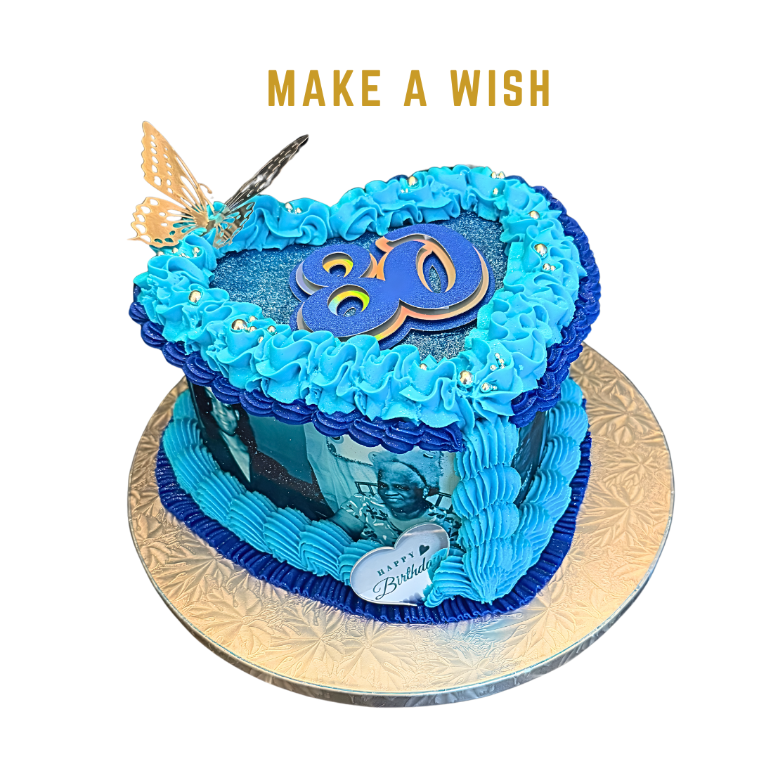 Blue heart-shaped birthday cake with a photograph of a woman on the side, a butterfly decoration on top, and a "Make a Wish" message. The cake has blue frosting and silver bead decorations, on a gold-colored base.