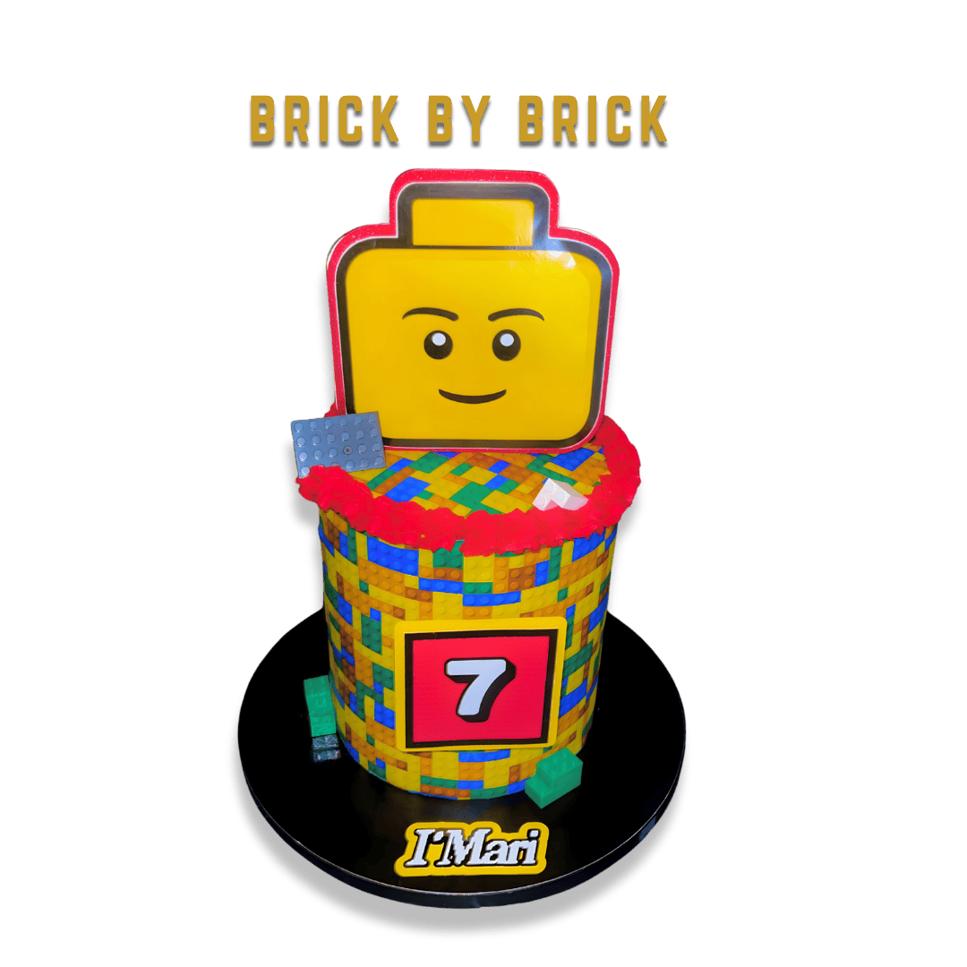 Lego-themed cake featuring a smiling Lego head and a brick pattern on the cake with the number 7, decorated with Lego bricks and the text "BRICK BY BRICK" and "I Mari".