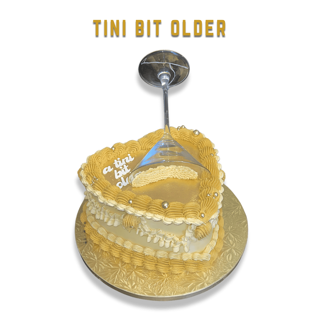 A heart-shaped cake with white and yellow frosting, silver edible beads, and a champagne glass on top, with the text "Tini Bit Older" above.