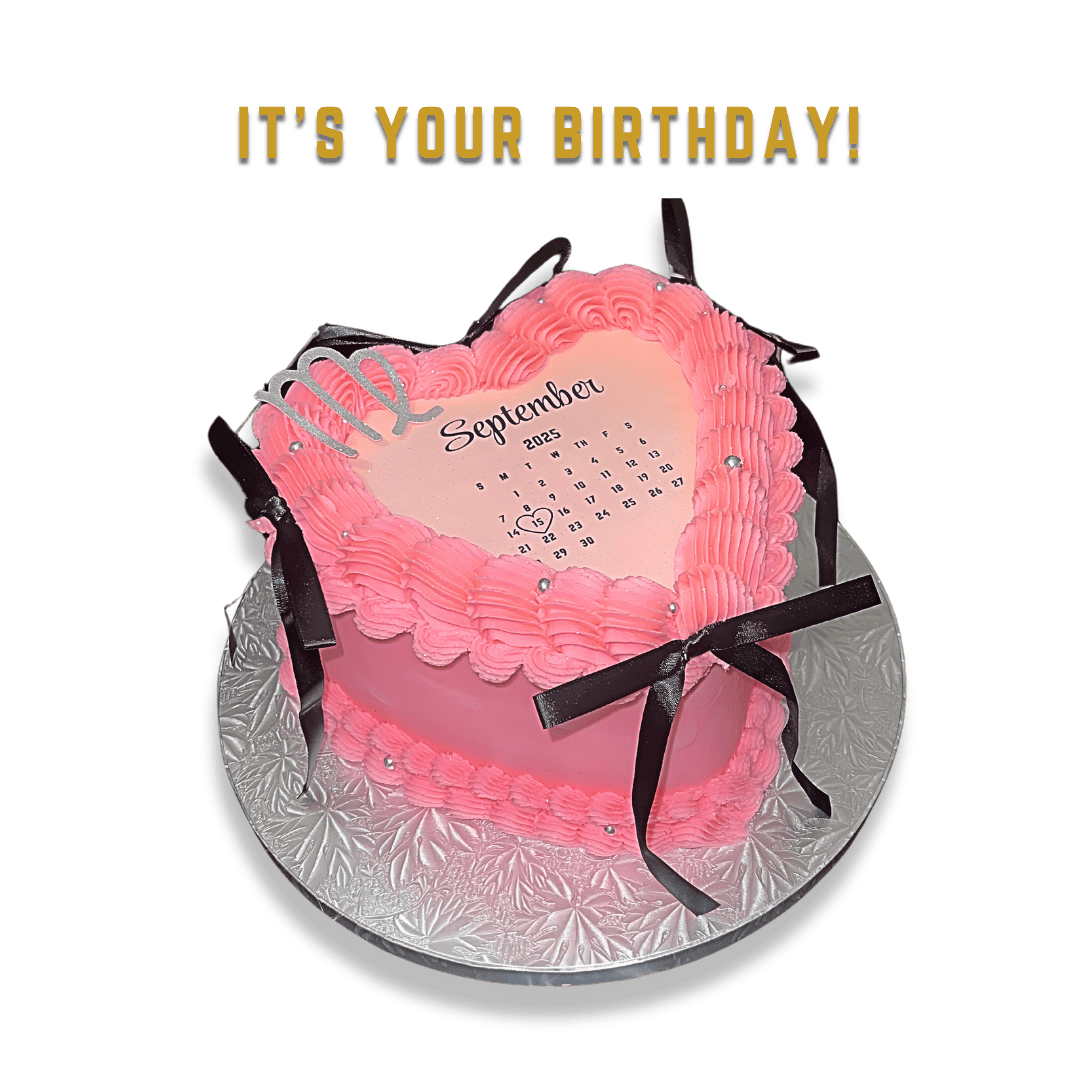 A pink heart-shaped birthday cake with piped frosting, black ribbons, and small silver decorations on a silver patterned cake board. The cake has a calendar for September 2025 on top and a silver 