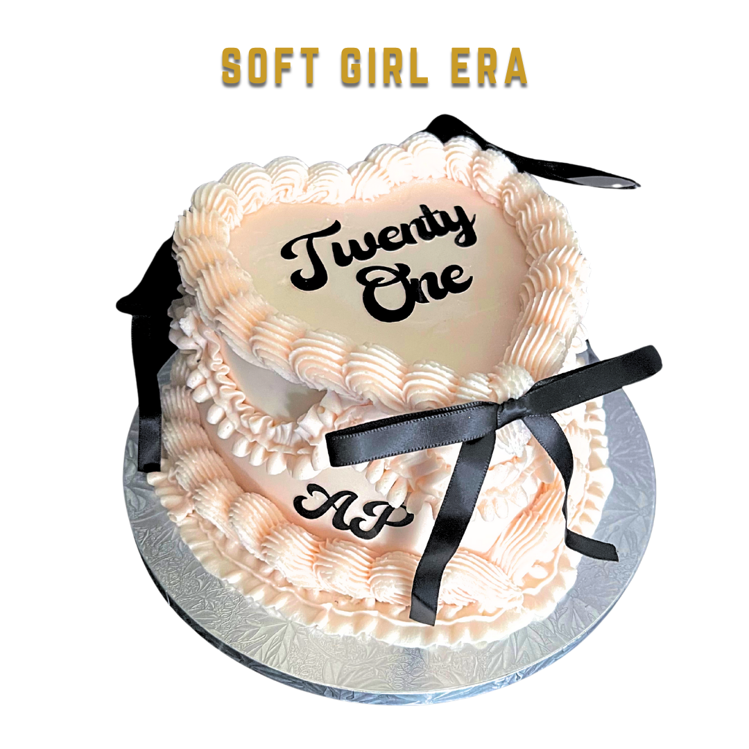 A heart-shaped birthday cake with pink and white frosting, a black ribbon bow, and the words 'Twenty One' and 'AP' written on it, celebrating a 21st birthday.