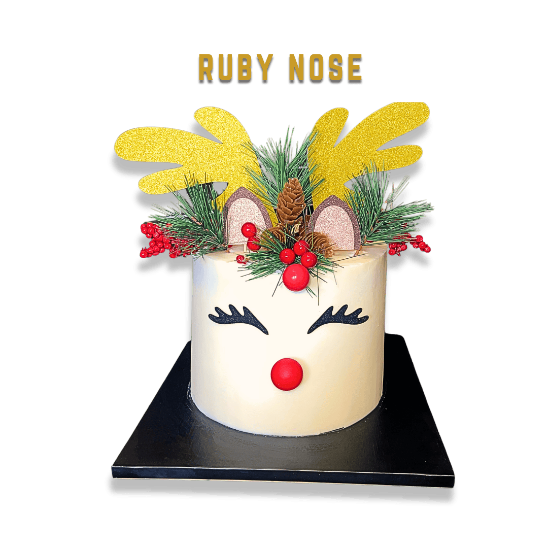 A holiday cake decorated as a reindeer with a white icing face, black eyelash-like details for closed eyes, a red ball for a nose, and reindeer antlers made of yellow glitter material, topped with pine branches, red berries, pinecones, and decorative