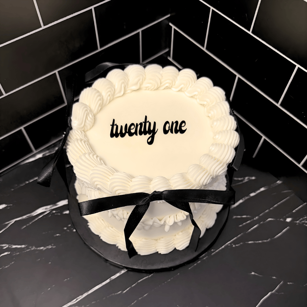 A heart-shaped cake decorated with yellow and white frosting, featuring a tilt glass on top, with a reflection creating a mirrored effect on the cake, and a message in white text in a foreign language on the side.