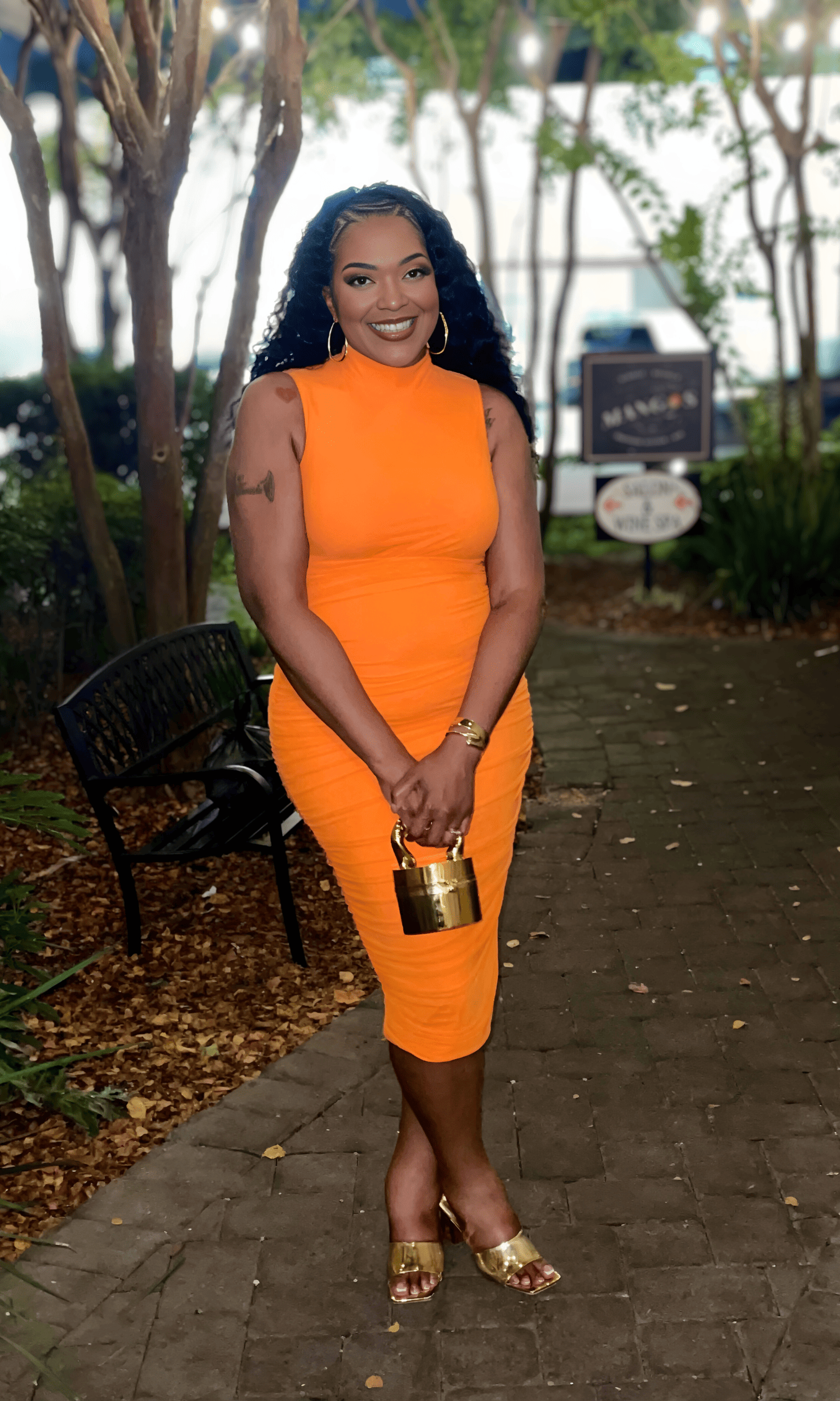 A woman standing outdoors on a brick pathway, wearing an orange fitted dress, gold heels, and holding a gold purse. She has curly black hair, gold hoop earrings, and is smiling at the camera. Trees and signs are in the background.