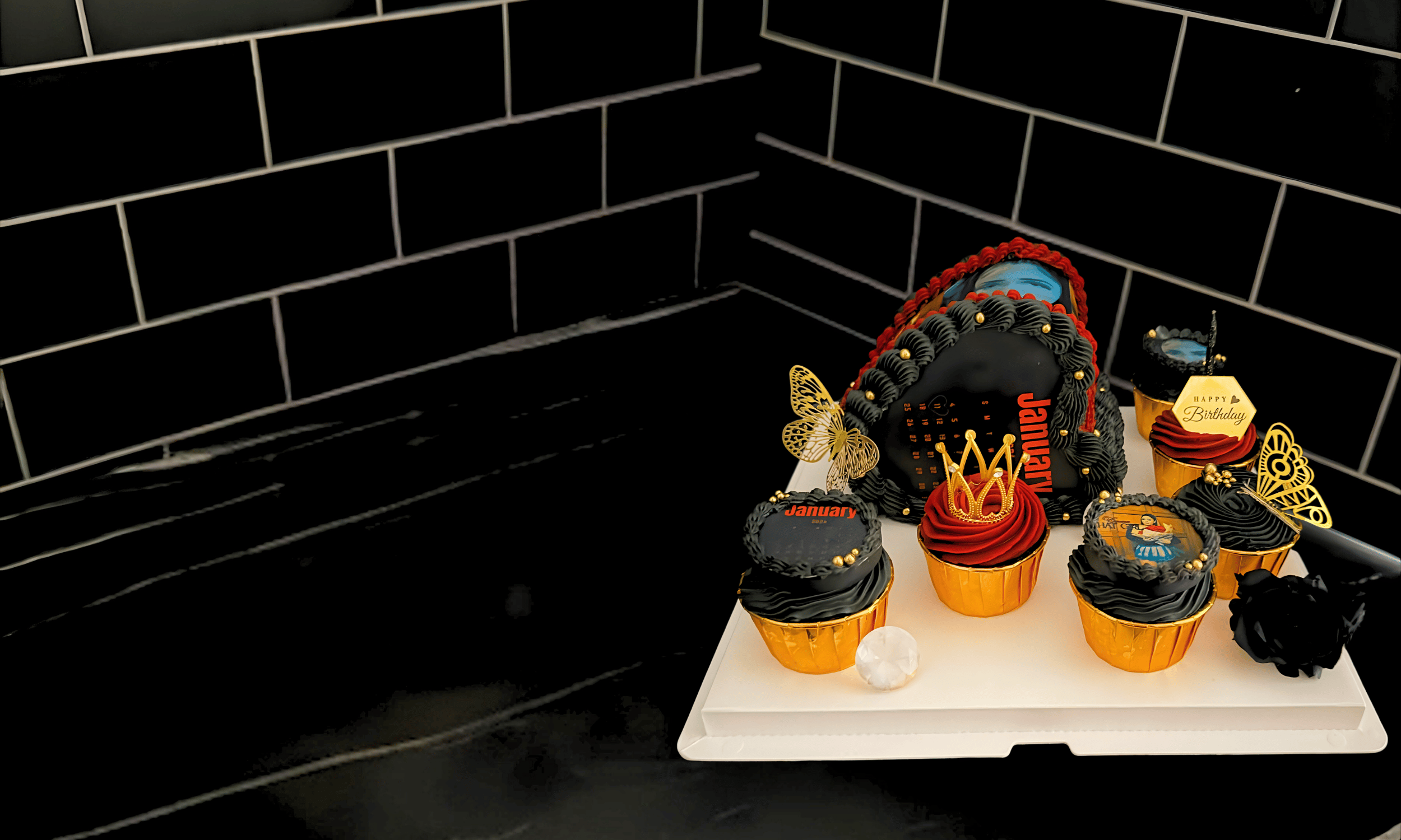 A birthday cake decorated with a black and red theme, surrounded by cupcakes with similar designs, some with 'January' and 'Happy Birthday' decorations.