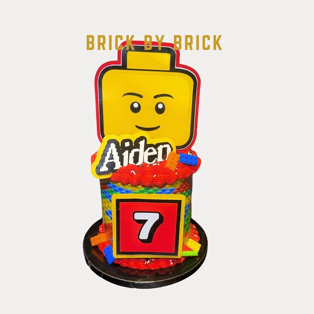 Lego-themed cake featuring a smiling Lego head and a brick pattern on the cake with the number 7, decorated with Lego bricks and the text "BRICK BY BRICK".