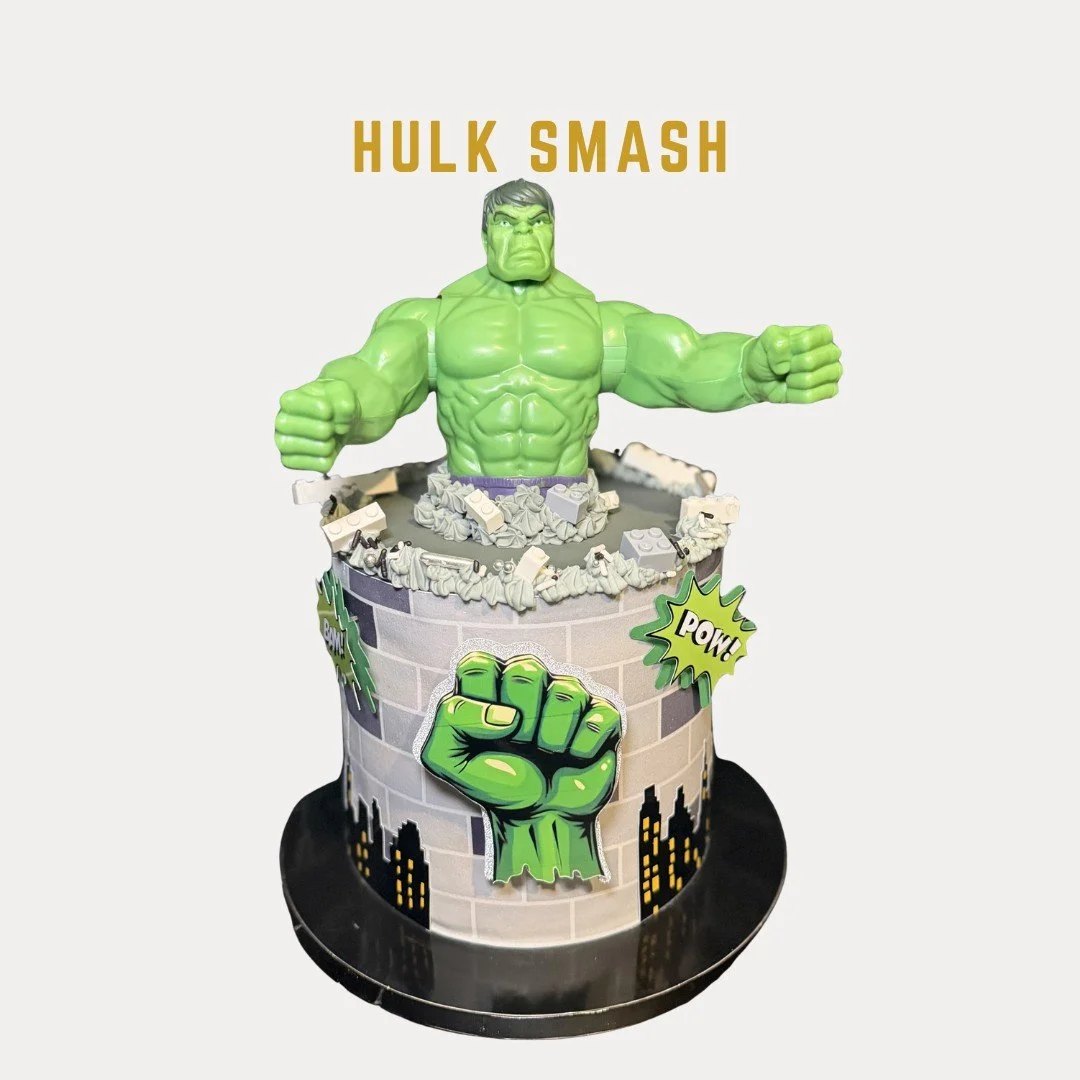 Hulk Smash Cake is Hulk-themed birthday cake featuring a large Hulk figure breaking through a brick wall with comic book style decorations including words like 'Pow!' and 'Bam!' and a fist symbol.