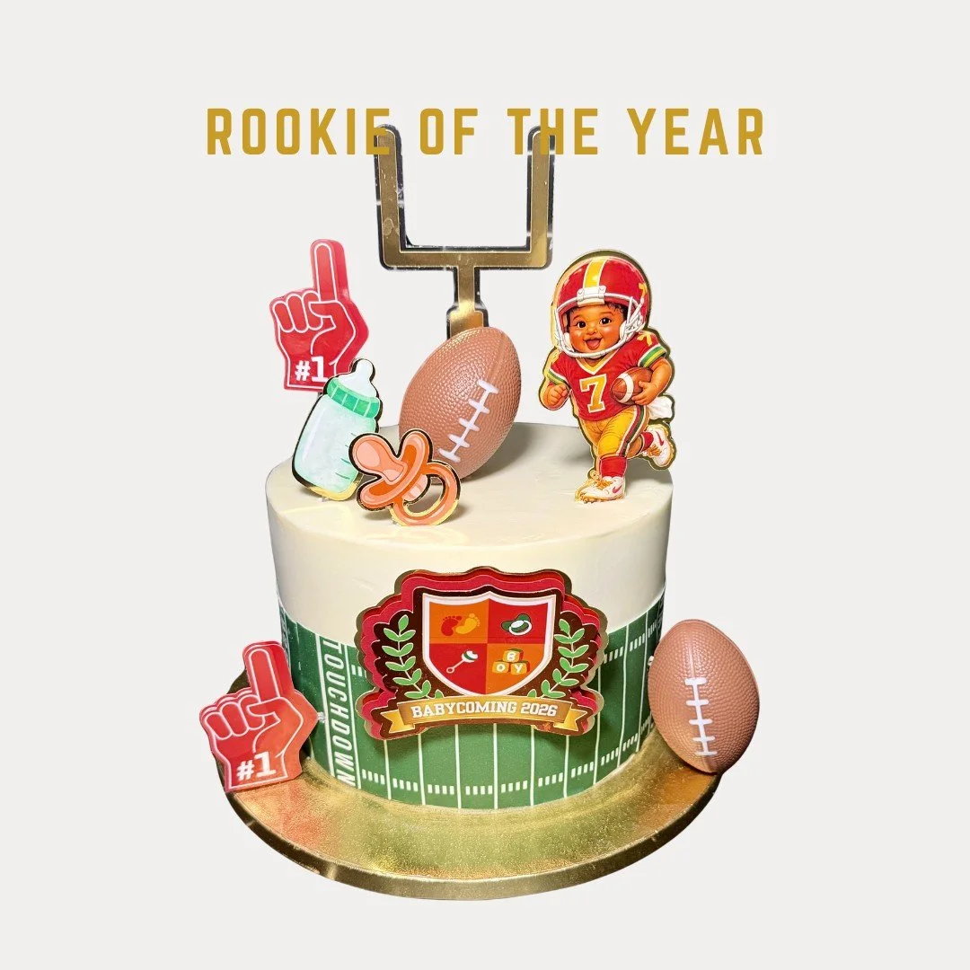 Rookie of the year is a cake decorated with football theme, featuring a football player cartoon, football confetti, baby bottles, and foam fingers, with text 'Rookie of the Year' and a badge that says 'Baby Coming 2026'.