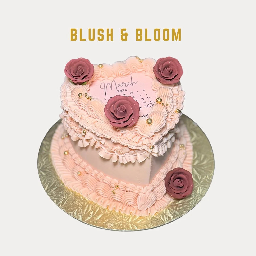 Heart-shaped pink cake decorated with pink and burgundy roses, gold beads, and pink icing, with a calendar and 'March 2026' written on top, on a gold cake board. The text 'BLUSH & BLOOM' is above the cake.