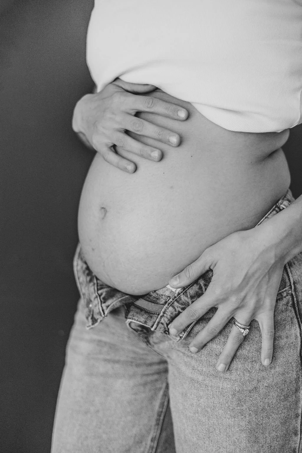 Close-up of a pregnant woman with her hands on her belly, wearing a white top and jeans, in black and white.