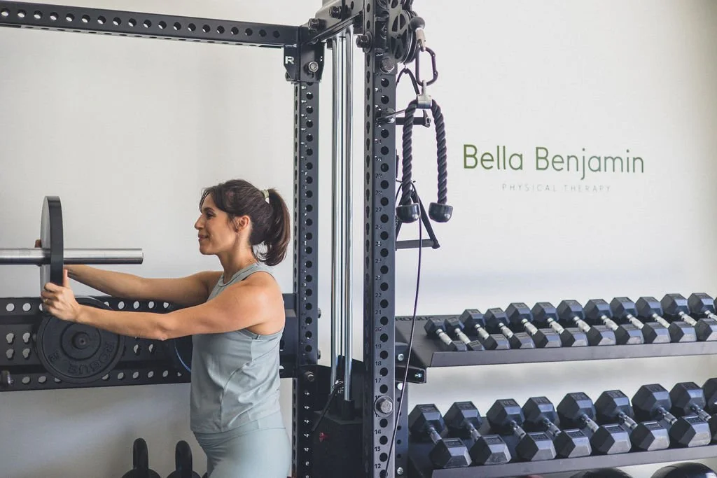 A woman in athletic wear exercising with a weight plate on a barbell in a gym, with a rack of dumbbells in the background and gym equipment hanging from a strong metal frame.