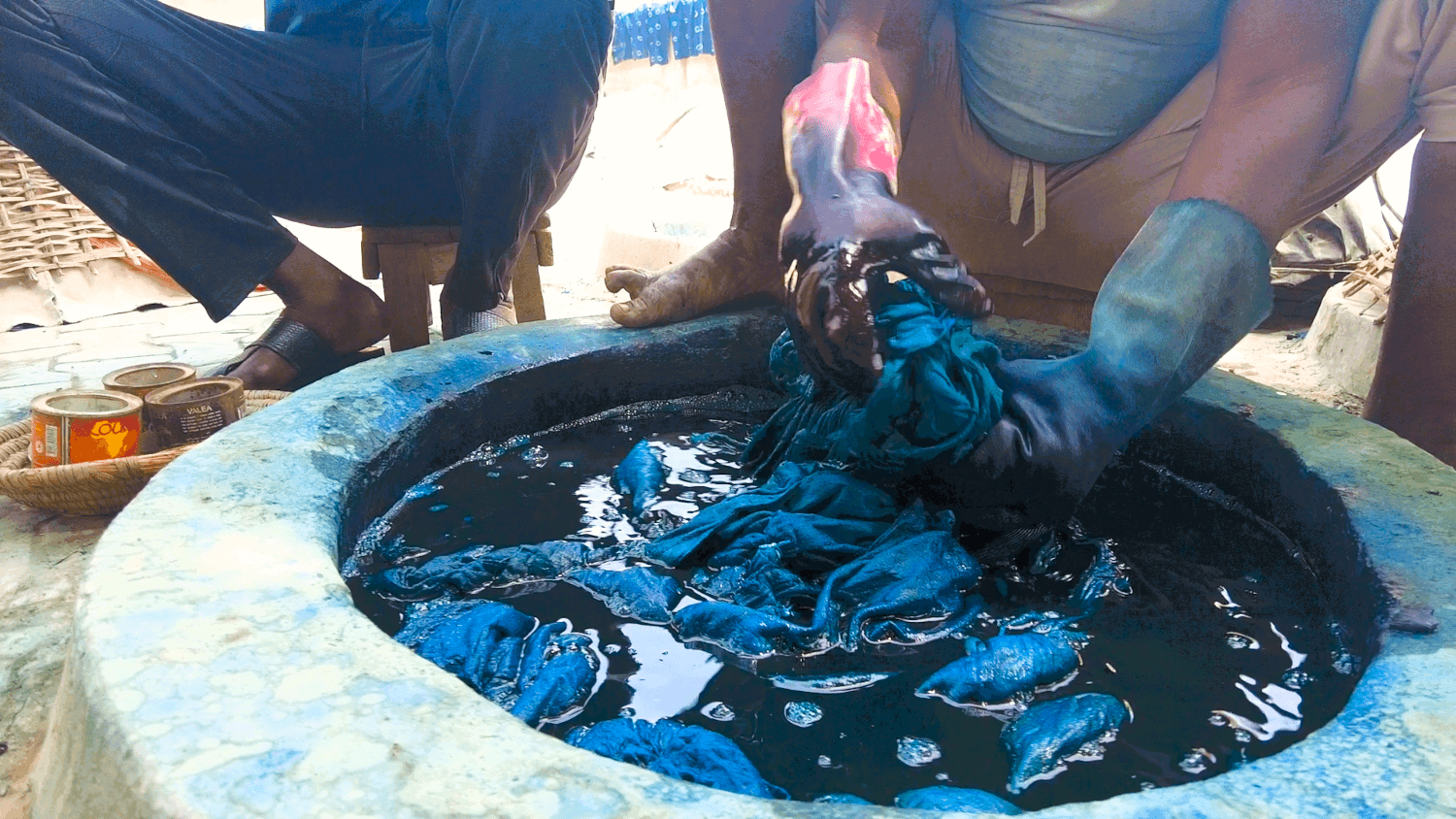 Hands working indigo-dyed fabric in a circular dye pit at Kofar Mata in Kano, Nigeria.