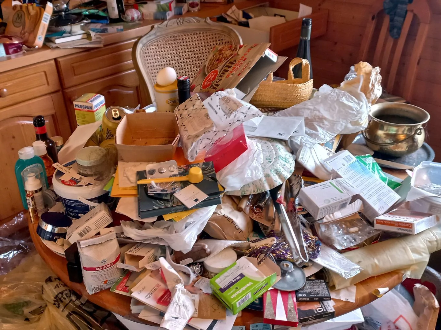Cluttered dining table piled with papers, containers, household items, and miscellaneous objects in a hoarded interior space