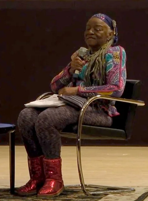 Artist Faith Ringgold seated onstage holding a microphone while speaking during a public symposium at the Brooklyn Museum.