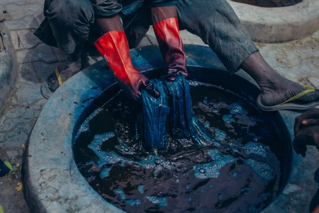 A Nigerian indigo dyer wearing protective gloves works blue cloth by hand in a large circular dye pit filled with dark indigo liquid.