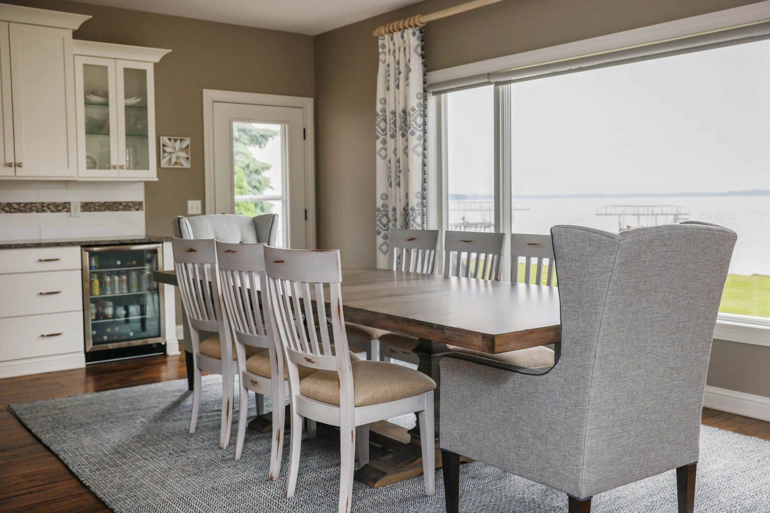 Dining room with large window, and lightly patterned white drapes. Large dining table with rustic white chairs and lager padded chairs at the heads of the table. 