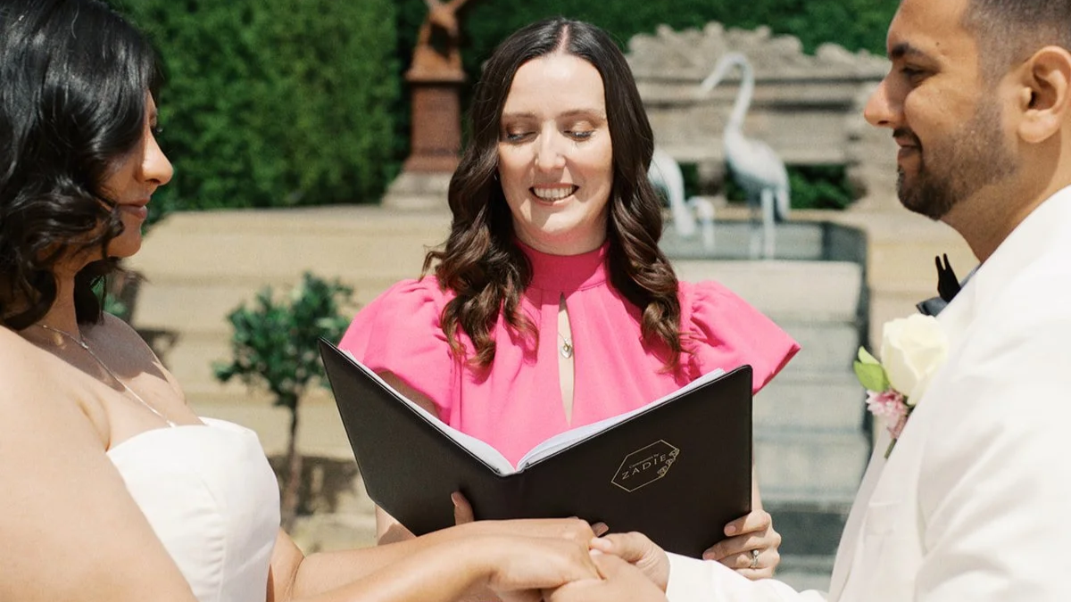 Wedding celebrant in pink dress reading ceremony from folder with wedding couple