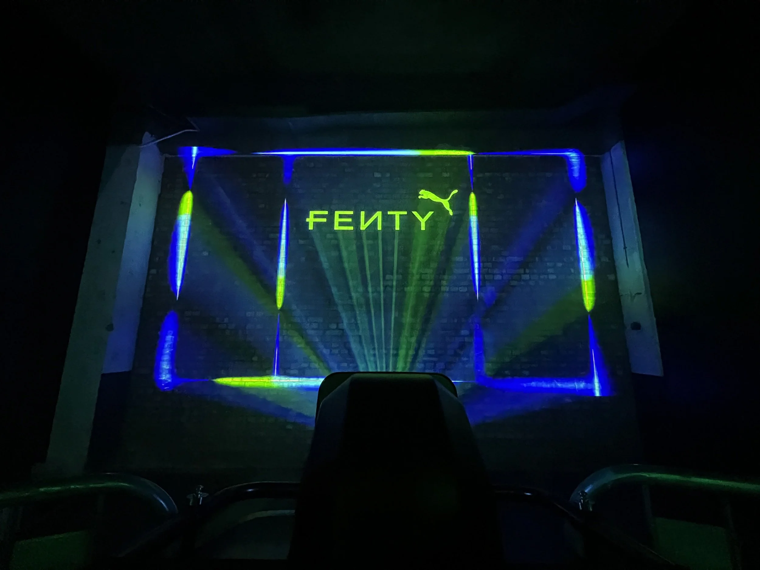 Projection mapping displaying the Fenty Puma logo on a brick wall with blue and green light beams