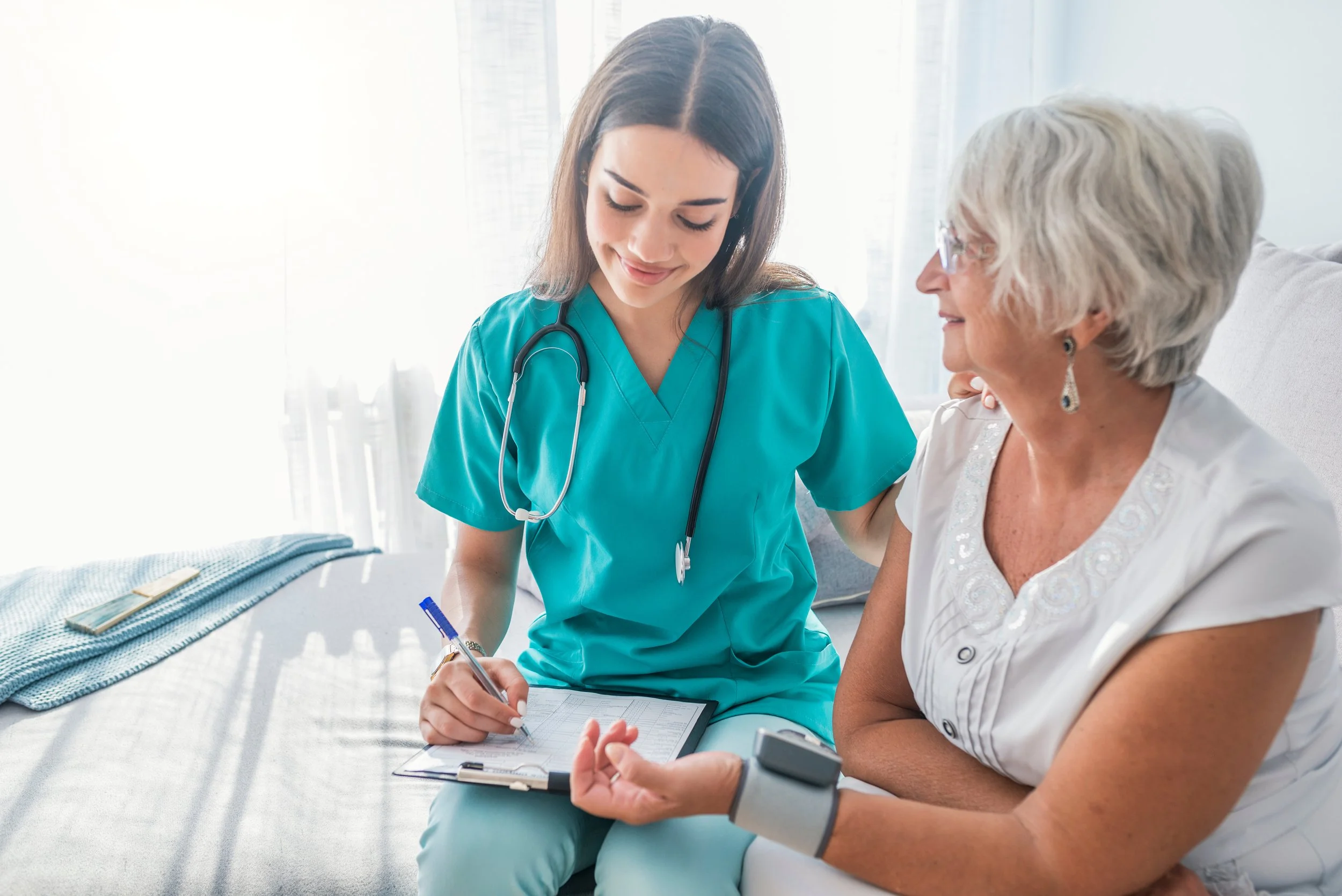 Nurse in teal scrubs measuring blood pressure of an elderly woman at home, writing on a clipboard.