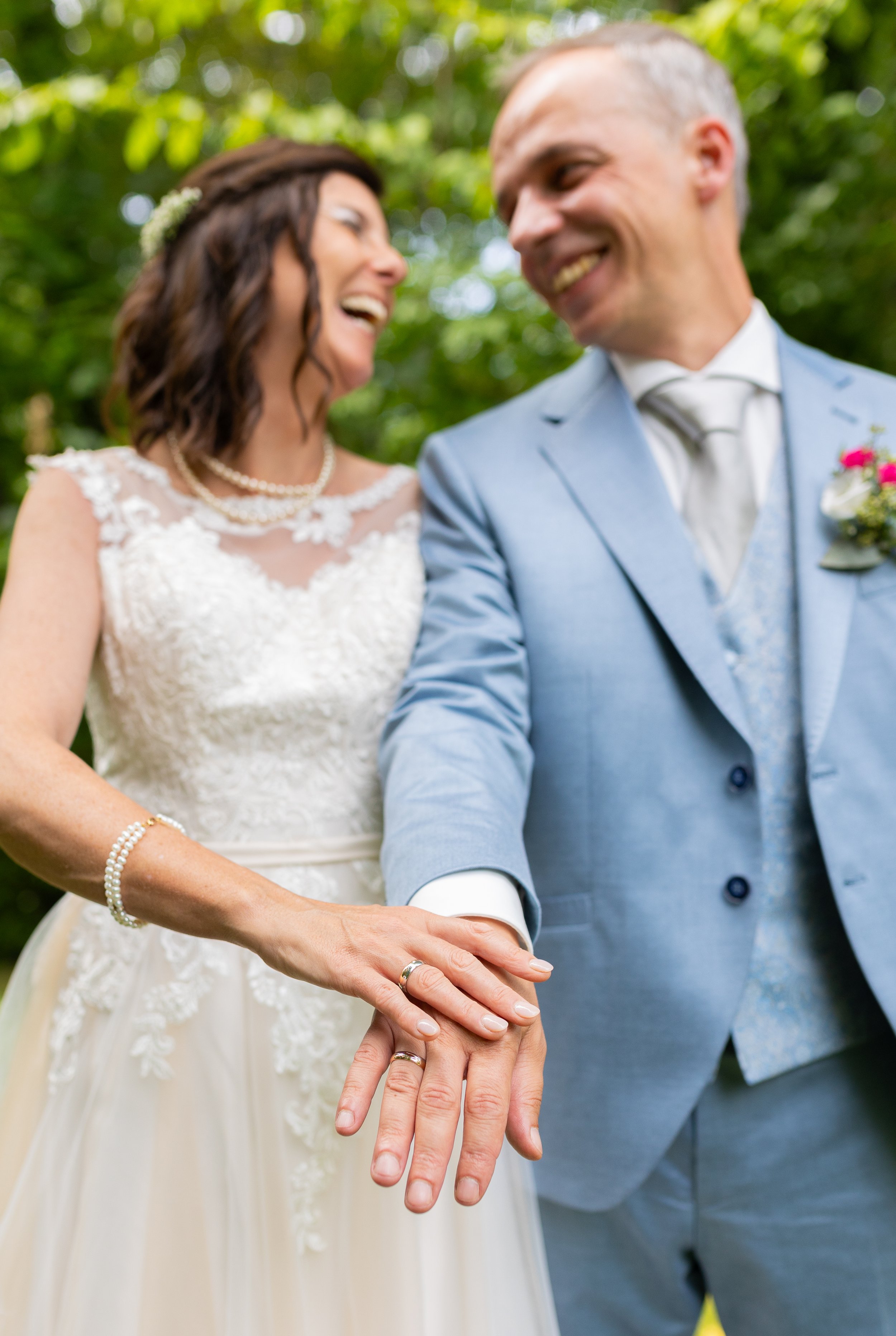 Braut und Bräutigam in Hochzeitskleidung, lächelnd, mit ineinandergelegten Händen und Eheringen.