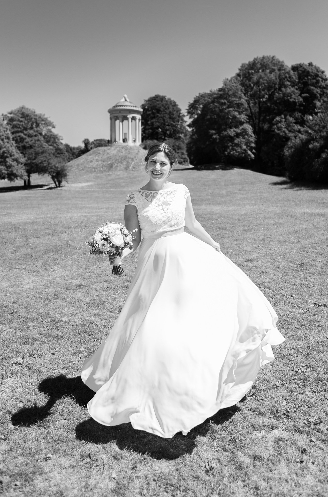 Braut in einem Hochzeitskleid hält einen Blumenstrauß, stehend auf einer Wiese mit einem Tempel im Hintergrund.